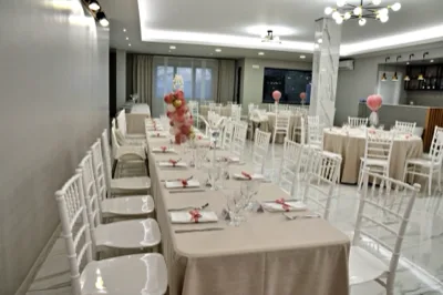 Sala apparecchiata per battesimo con una lunga tavolata centrale, tavoli rotondi sullo sfondo, sedie bianche e dettagli rosa nei nastri dei tovaglioli e nelle decorazioni. L'ambiente è luminoso e ordinato, con un allestimento delicato e coerente.