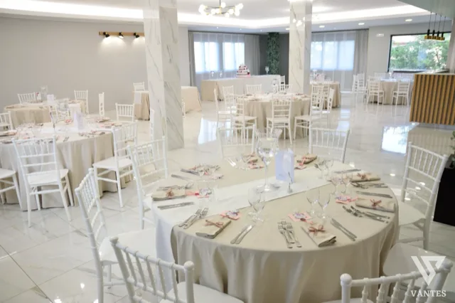 Sala per ricevimento elegante con tavoli rotondi apparecchiati, dettagli rosa cipria e tavolo della torta sul fondo. L'ambiente è luminoso, raffinato e adatto a matrimonio o cerimonia formale.