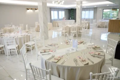 Sala per ricevimento elegante con tavoli rotondi apparecchiati, dettagli rosa cipria e tavolo della torta sul fondo. L'ambiente è luminoso, raffinato e adatto a matrimonio o cerimonia formale.