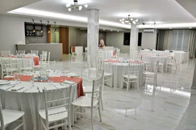 Sala ricevimenti interna con tavoli rotondi apparecchiati, sedie bianche, runner rosa salmone e ampio pavimento lucido effetto marmo. Sullo sfondo si vedono il bancone bar e i pilastri chiari che definiscono uno spazio moderno ed elegante.