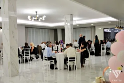 Sala ricevimenti con ospiti seduti ai tavoli durante un pranzo o una cena di cerimonia, in un ambiente luminoso con pavimento effetto marmo e sedie bianche. Sul lato destro si intravedono anche decorazioni con palloncini che rafforzano il contesto festivo.