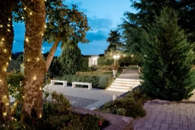 Ingresso serale del giardino con gradini illuminati, vialetto in pietra, alberi decorati con luci e vegetazione curata ai lati. L'atmosfera è elegante e accogliente, ideale per l'arrivo degli ospiti in location.