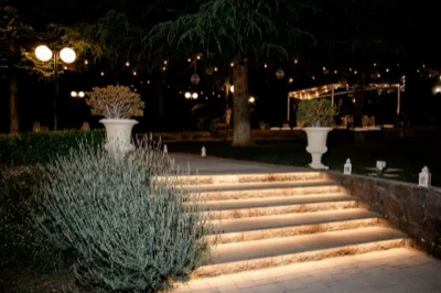Scalinata serale del giardino con gradini illuminati, lanterne basse e luci sospese tra gli alberi. L'inquadratura ravvicinata valorizza l'atmosfera scenografica dell'area esterna di notte.