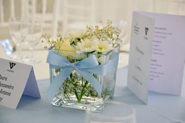 Dettaglio di un centrotavola in vetro con fiori bianchi e nastro azzurro, affiancato da segnaposto e menu stampati su un tavolo apparecchiato per battesimo. L'immagine mette in risalto la cura dei dettagli e la palette chiara della mise en place.
