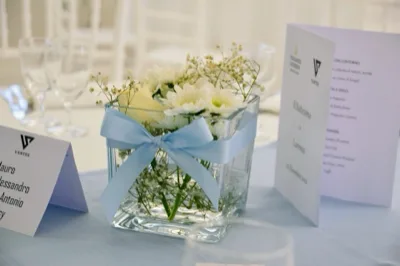 Dettaglio di un centrotavola in vetro con fiori bianchi e nastro azzurro, affiancato da segnaposto e menu stampati su un tavolo apparecchiato per battesimo. L'immagine mette in risalto la cura dei dettagli e la palette chiara della mise en place.
