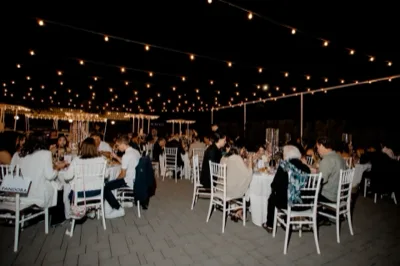 Ricevimento catering all'aperto con ospiti seduti a tavoli rotondi sotto una rete di luci sospese. La scena mostra una cena serale elegante e conviviale in uno spazio esterno ampio e curato.