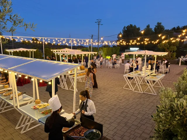 Ampia vista serale dello spiazzale esterno con isole buffet illuminate, gazebo bianchi, catenarie luminose e ospiti distribuiti nell'area evento. In primo piano si vedono staff di cucina e servizio mentre preparano il buffet all'aperto.