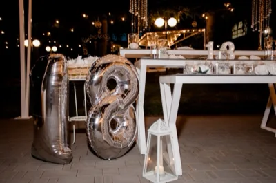 Scenografia per un diciottesimo compleanno in esterno con grandi numeri 18 argentati, tavolo bianco decorato, candele e luci sospese sullo sfondo. L'allestimento è elegante e pensato come punto focale della festa serale.