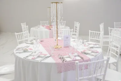 Tavoli rotondi apparecchiati in sala con runner rosa, sedie bianche e alti centrotavola dorati con pendenti di cristallo. La mise en place è luminosa e ordinata, pensata per un battesimo o una cerimonia dai toni delicati.