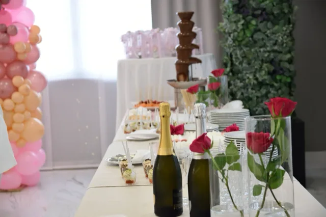 Tavolo dessert in sala con fontana di cioccolato, bottiglie di spumante, rose rosse in vasi cilindrici e dolci monoporzione disposti lungo il piano. Sul lato sinistro si intravede anche una colonna di palloncini rosa e pesca che completa l'allestimento del battesimo.