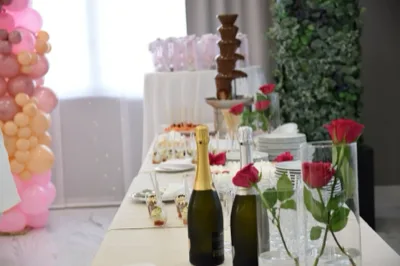 Tavolo dessert in sala con fontana di cioccolato, bottiglie di spumante, rose rosse in vasi cilindrici e dolci monoporzione disposti lungo il piano. Sul lato sinistro si intravede anche una colonna di palloncini rosa e pesca che completa l'allestimento del battesimo.