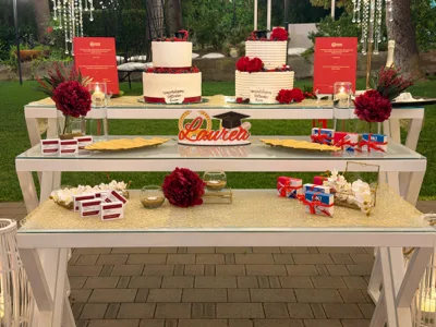 Tavolo torta per festa di laurea all'aperto con due torte a piani bianche, libretti rossi che richiamano la tesi, rose bordeaux, candele e bomboniere disposte su tre livelli. Al centro spicca la scritta decorativa 'Laurea', mentre sullo sfondo si vedono il prato, alberi e sedute del giardino illuminato di sera.