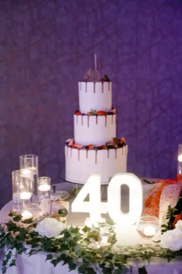 Torta scenografica per i 40 anni a tre piani, decorata con drip scuro, frutta fresca e numero luminoso 40 in primo piano. Candele e composizioni floreali completano l'allestimento elegante del tavolo torta.