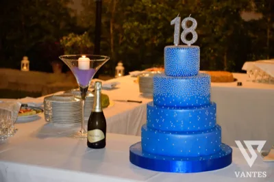 Torta blu a più piani per i 18 anni, decorata con perline argentate e topper con numero 18. Il tavolo è allestito in esterno con candela, bottiglia e piatti da servizio pronti per il taglio della torta.