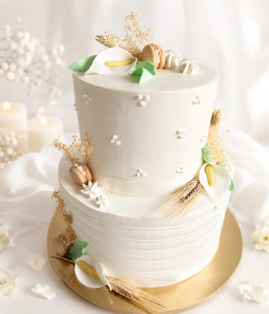 Torta bianca a due piani per Cresima con decorazioni di calle, spighe dorate, macarons chiari e piccoli dettagli in pasta di zucchero. L'inquadratura ravvicinata valorizza uno stile pulito ed elegante su sfondo chiaro e luminoso.