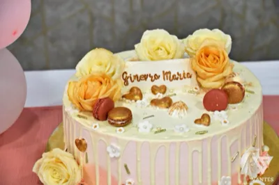 Primo piano di una torta rosa e avorio con colature chiare, rose decorative, macarons e piccoli cuori dorati sulla superficie. La composizione è elegante e delicata, pensata per un battesimo o una festa dai toni pastello.