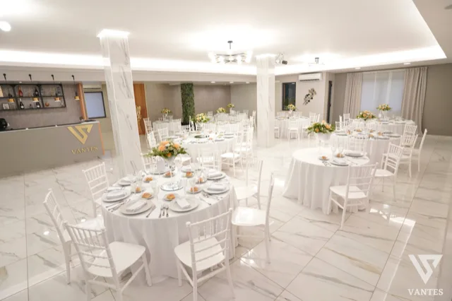Vista ampia della sala Vantes con tavoli rotondi apparecchiati, sedie bianche, centrotavola floreali chiari e bancone bar con logo in evidenza sulla sinistra. L'ambiente è luminoso, ordinato e rifinito con pavimento effetto marmo e linee moderne.