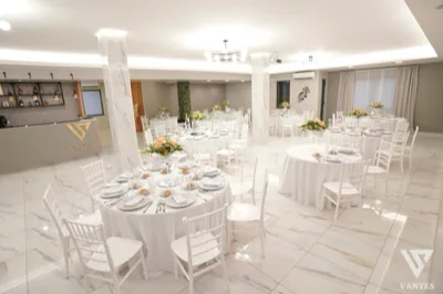 Vista ampia della sala Vantes con tavoli rotondi apparecchiati, sedie bianche, centrotavola floreali chiari e bancone bar con logo in evidenza sulla sinistra. L'ambiente è luminoso, ordinato e rifinito con pavimento effetto marmo e linee moderne.